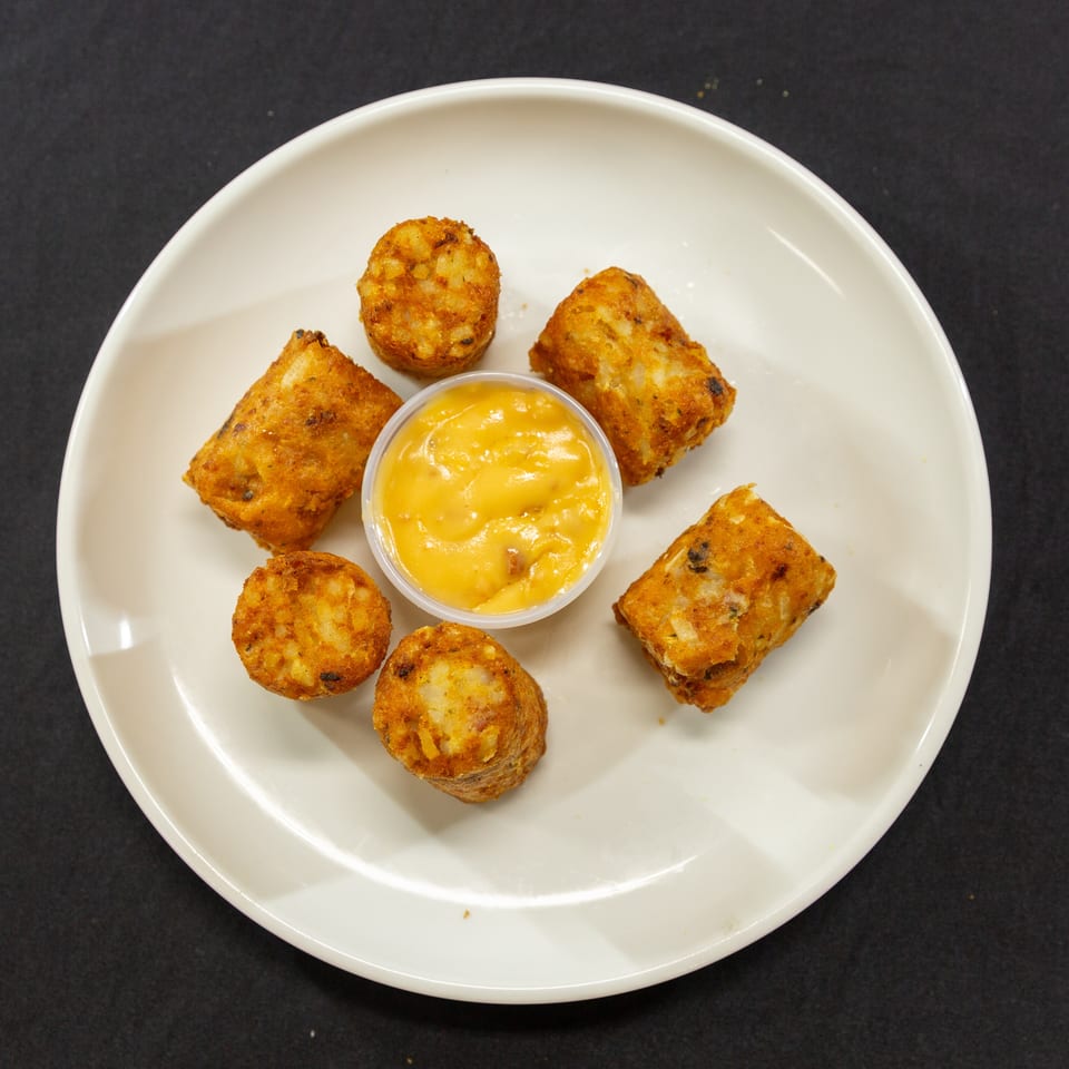 Golden-brown tater tots and cylindrical hash browns arranged around a cup of cheese sauce on a white plate.