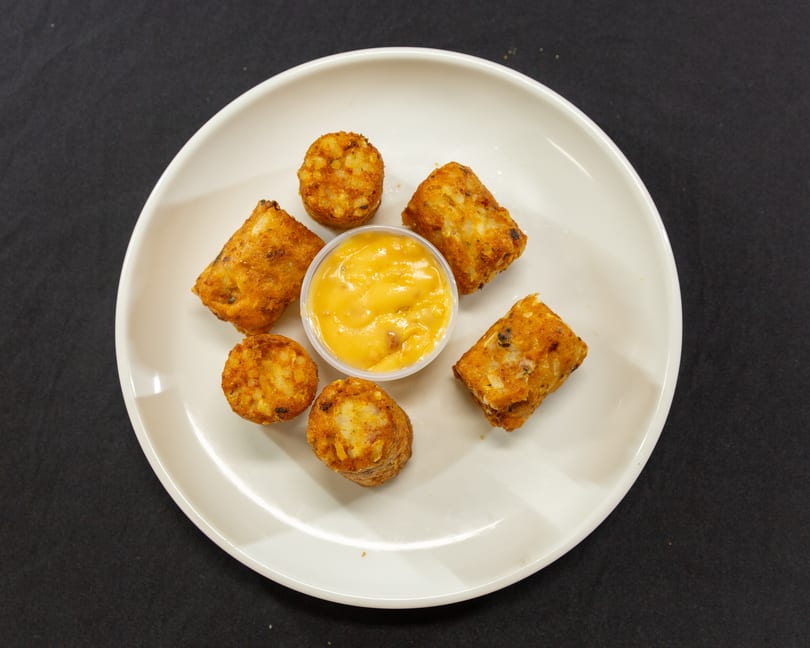 Golden-brown tater tots and cylindrical hash browns arranged around a cup of cheese sauce on a white plate.