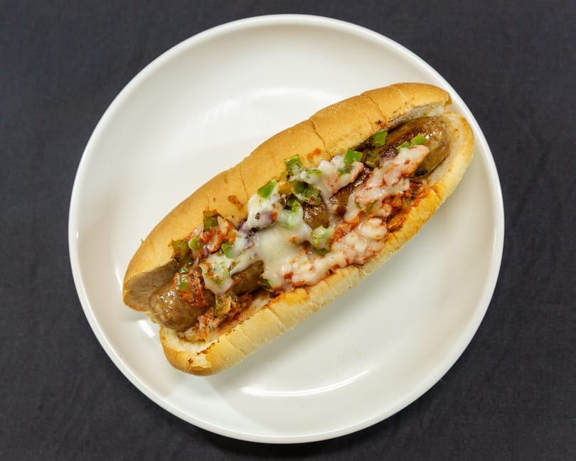 A sausage sandwich with melted cheese, green peppers, and red sauce on a white plate.