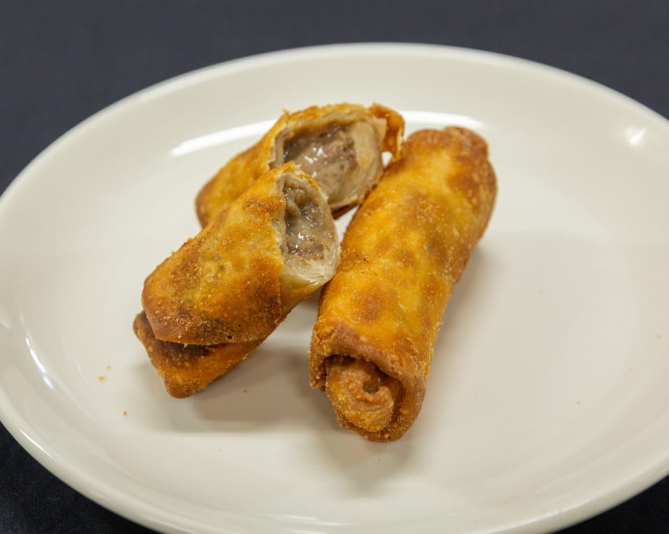 Three crispy fried egg rolls on a white plate, one cut open revealing a savory filling.