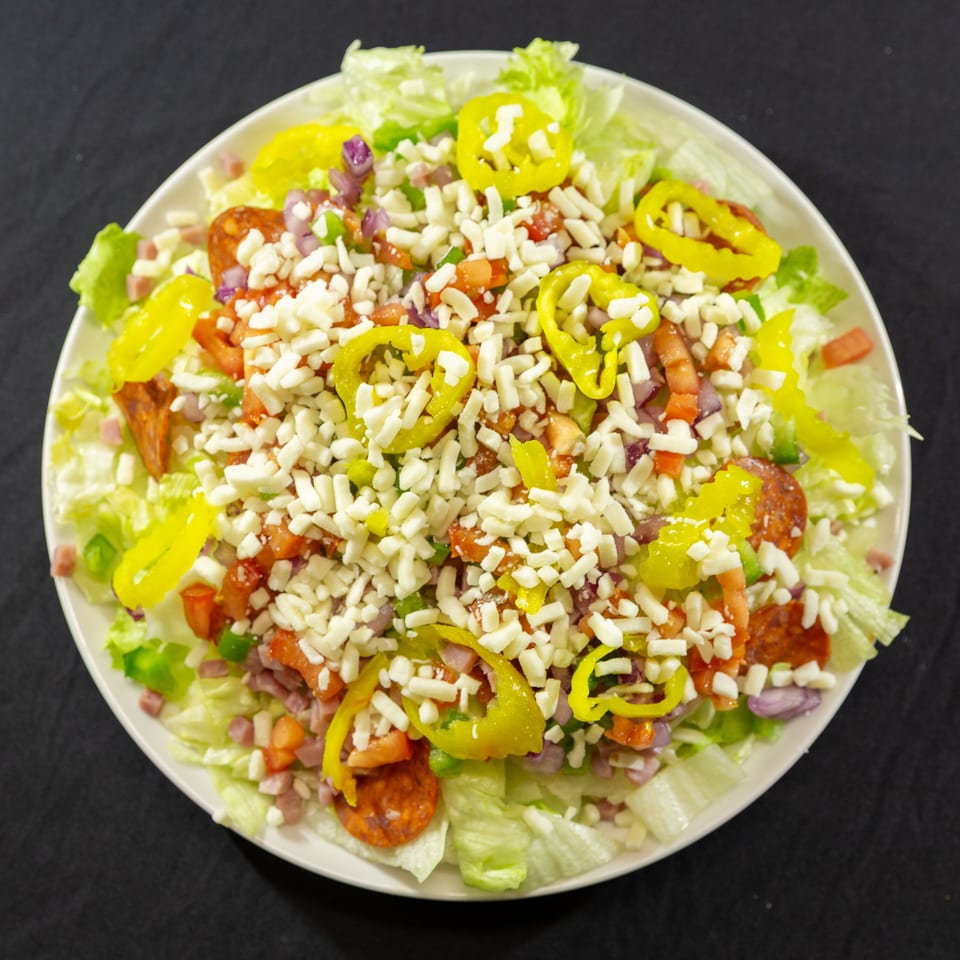 A large salad with lettuce, pepperoni, ham, tomatoes, onions, peppers, and shredded cheese.