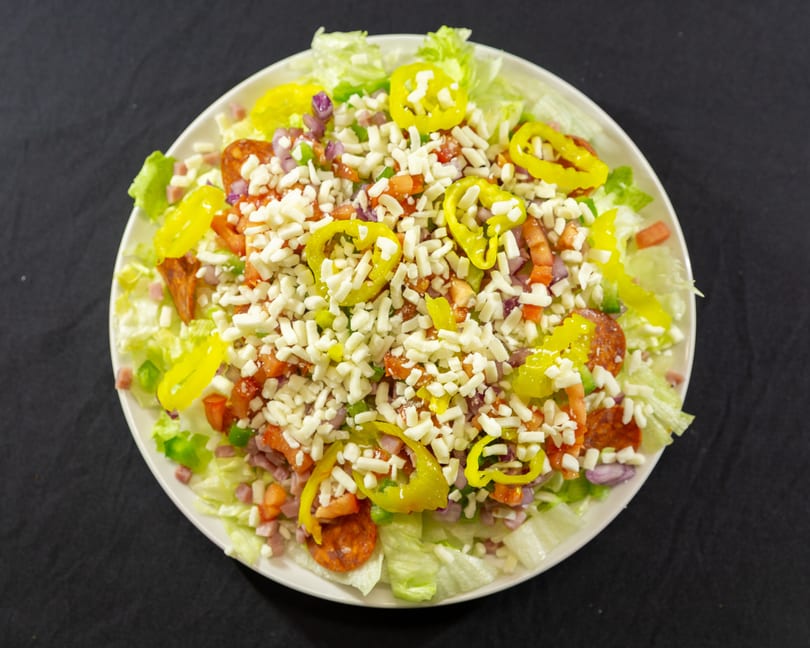 A large salad with lettuce, pepperoni, ham, tomatoes, onions, peppers, and shredded cheese.