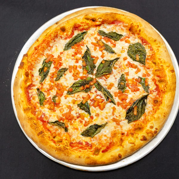 A whole Margherita pizza with fresh basil and diced tomatoes on a metal tray.