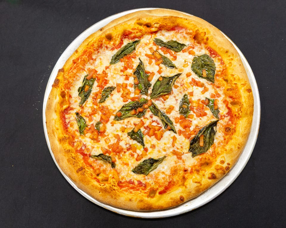 A whole Margherita pizza with fresh basil and diced tomatoes on a metal tray.